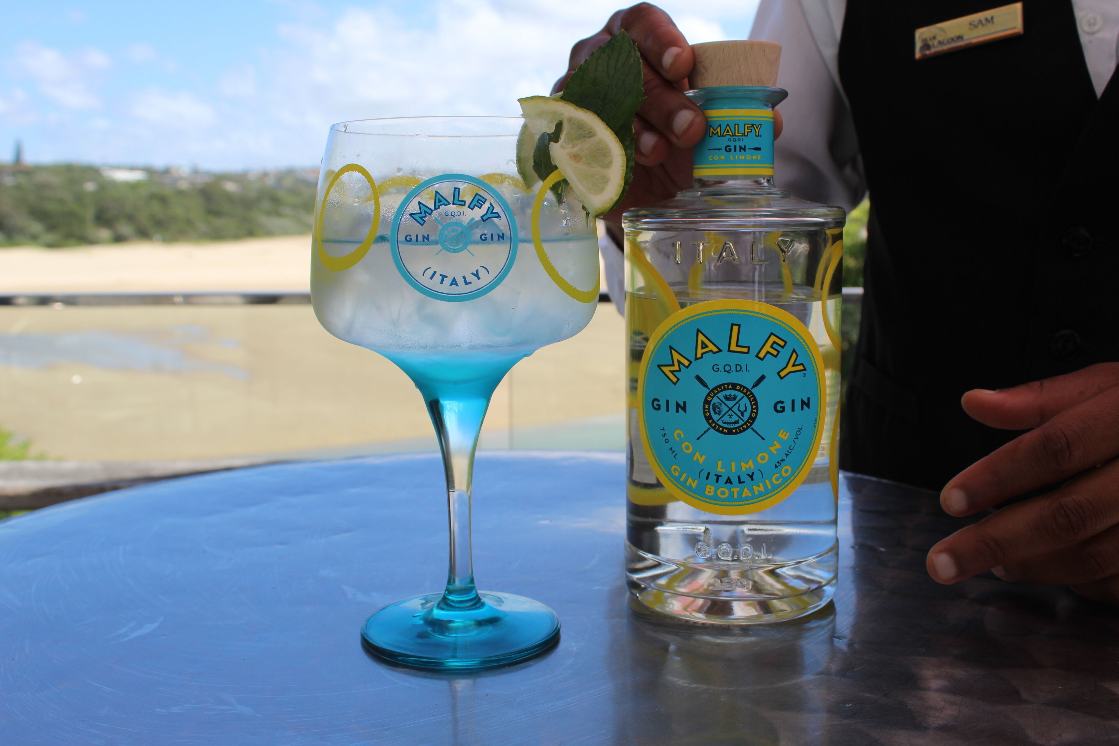 Uma garrafa de gin Malfy e um copo de gin Malfy com uma fatia de limão sobre uma mesa.