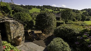 A stone building with a wooden bench and a table, surrounded by lush greenery and a view of rolling hills. - Townfoot Barn (Windermere)