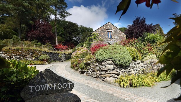 A stone building with a window, surrounded by a garden with various plants and a pathway leading to it. - Townfoot Barn (Windermere)