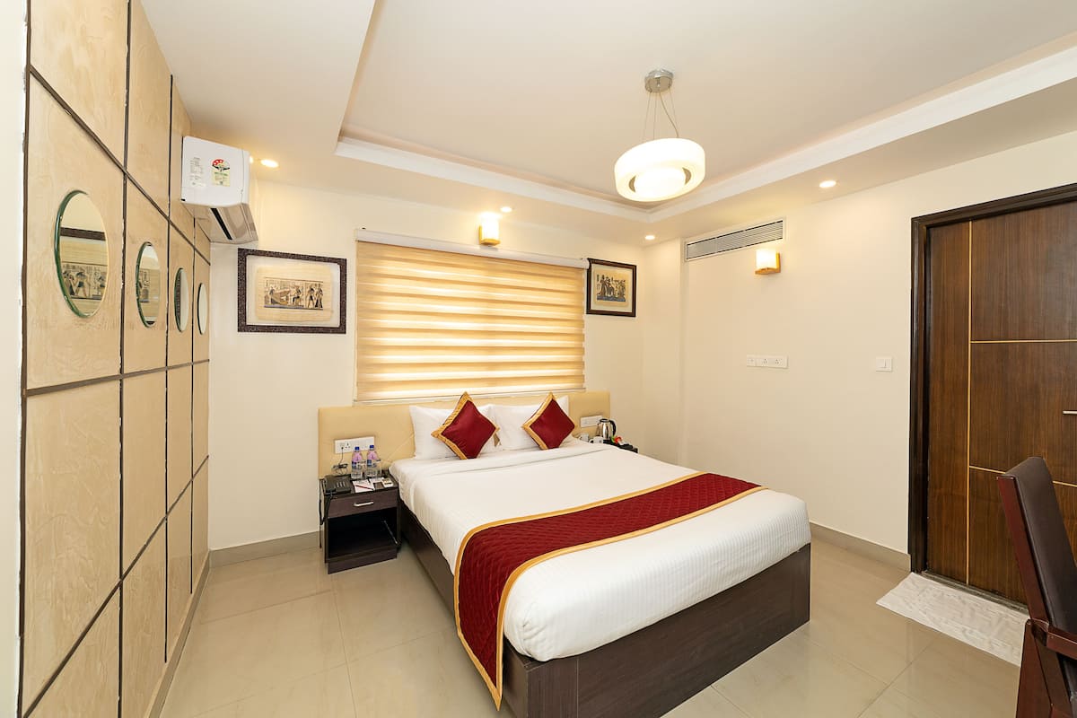 A hotel room with a large bed, a bedside table, a nightstand, a wardrobe, a mirror, a window with blinds, and a ceiling light fixture.