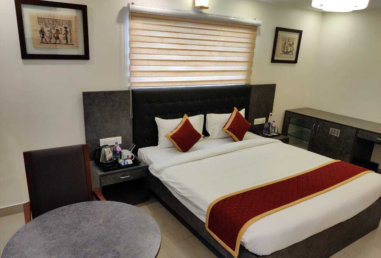 A hotel room with a bed, a bedside table with a lamp, a chair, a round table, and a window with blinds.