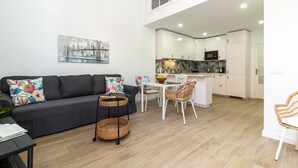 Una sala de estar moderna con un sofá, una mesa de comedor y sillas. Hay una zona de cocina con armarios blancos y un microondas.