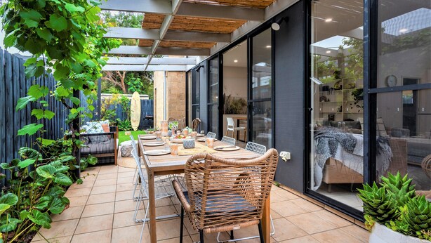 Une terrasse extérieure avec une table à manger pour quatre personnes, entourée de chaises en osier, d’une pergola en bois et de plantes en pot.