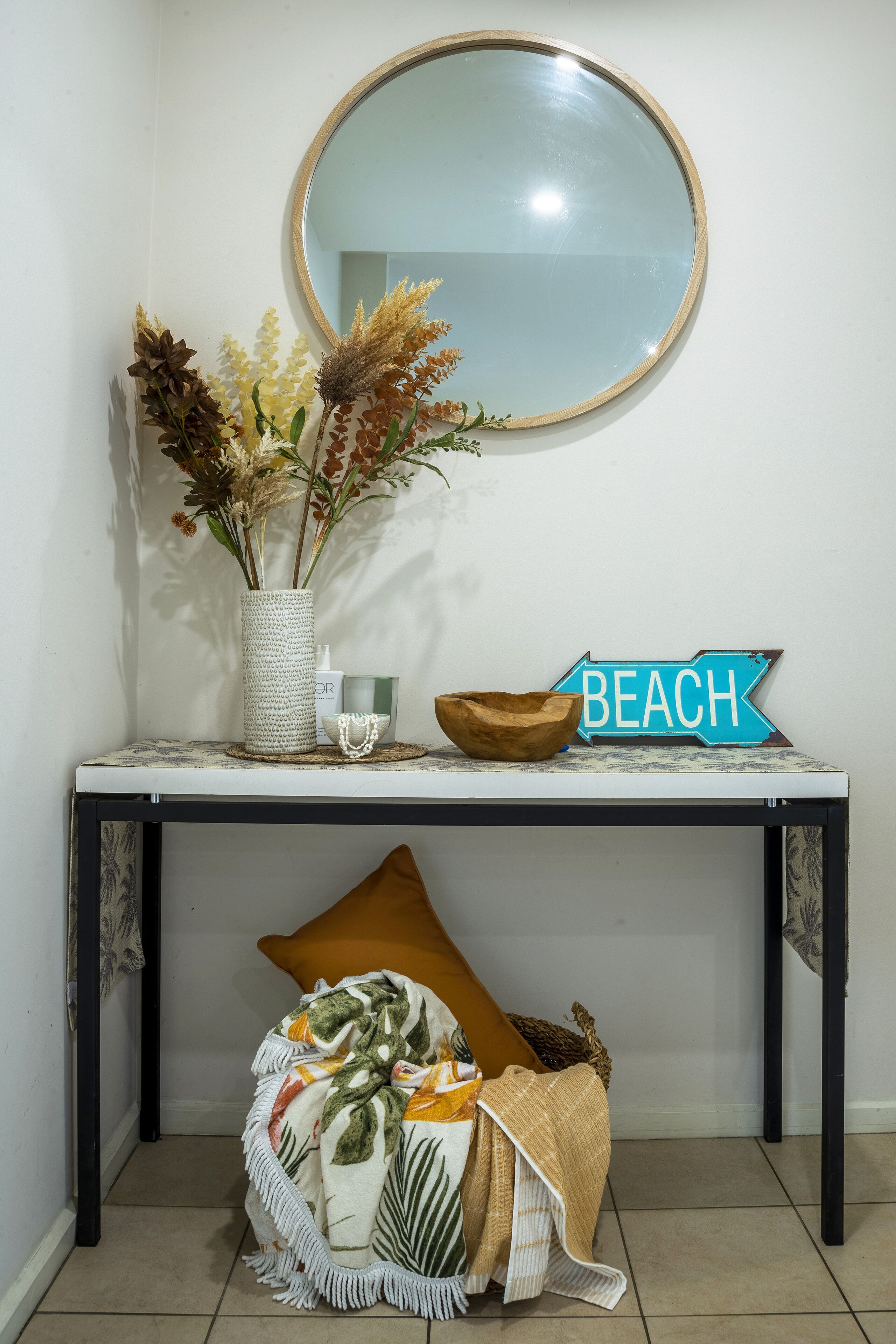 Un tavolo consolle con uno specchio, un vaso con fiori secchi, una ciotola e un cartello con la scritta 'BEACH'.