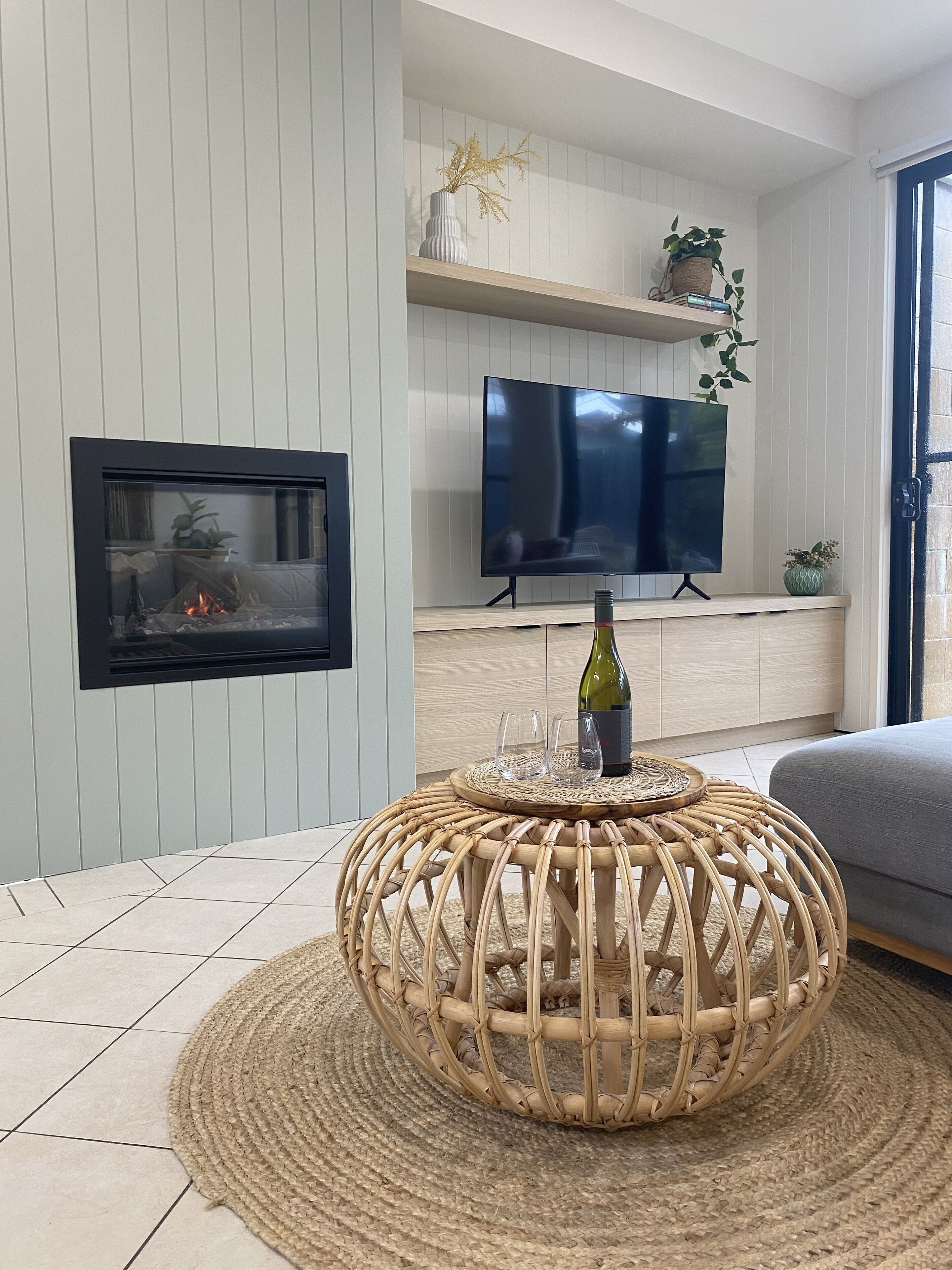 A modern living room with a fireplace, a flat-screen TV, and a wicker coffee table with a bottle and glasses.