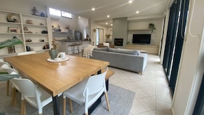 Un salon moderne avec une table à manger en bois, des chaises blanches, un canapé gris et un téléviseur fixé au mur.