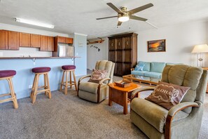 Ein Wohnzimmer mit einer Couch, zwei Sesseln, einem Couchtisch und einer Küchenzeile mit Holdekor und einem Kühlschrank.
