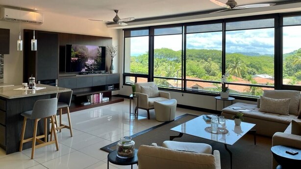 Una sala de estar moderna con un televisor grande, asientos cómodos, una zona de cocina y vistas a zonas verdes a través de amplios ventanales.