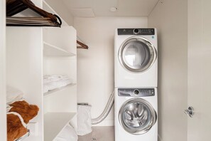 A laundry room with a stacked washer and dryer, a drying rack, and a white shelving unit.