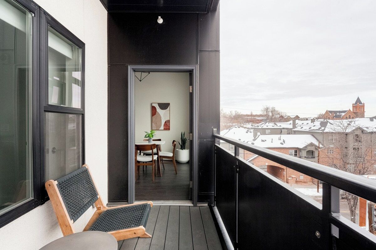 A balcony with a view of snow-covered buildings, a wooden chair, and a table with chairs.