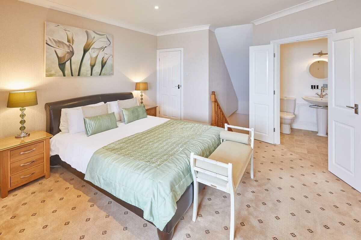 A bedroom with a bed, bedside tables, a nightstand, a chair, a painting, a door, and a bathroom visible through an open door.