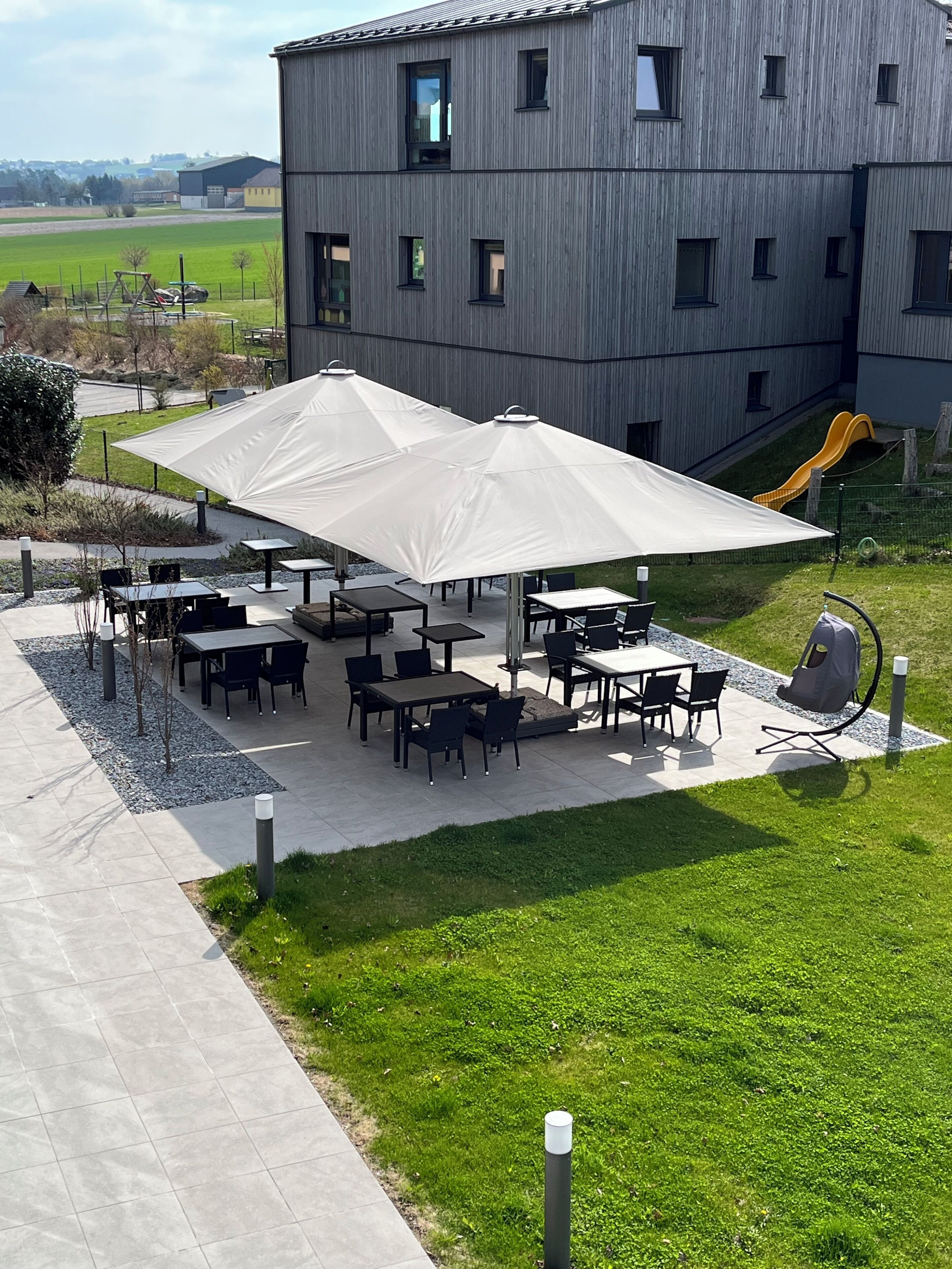 An outdoor patio area with tables and chairs, a large umbrella, and a playground with a slide.