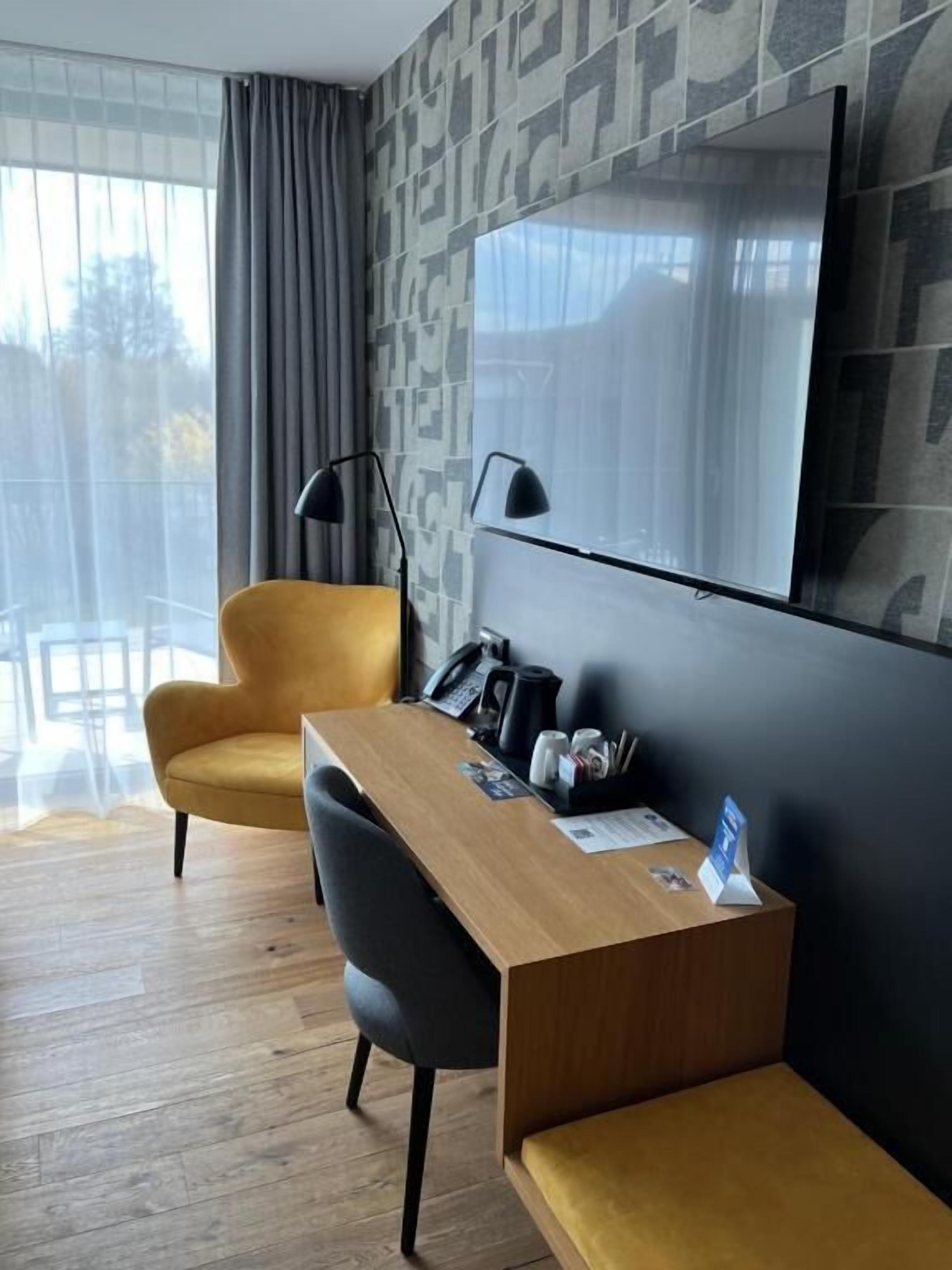 A modern hotel room with a wooden desk, a black chair, a yellow sofa, a large mirror, and a patterned wall.