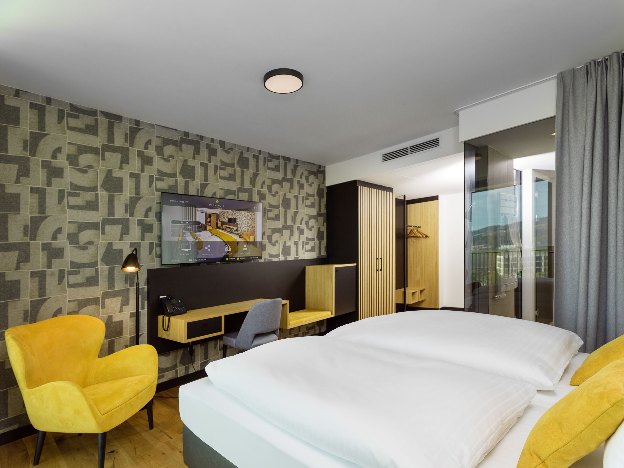 A modern hotel room with a large bed, a TV mounted on the wall, a desk, and a wardrobe.