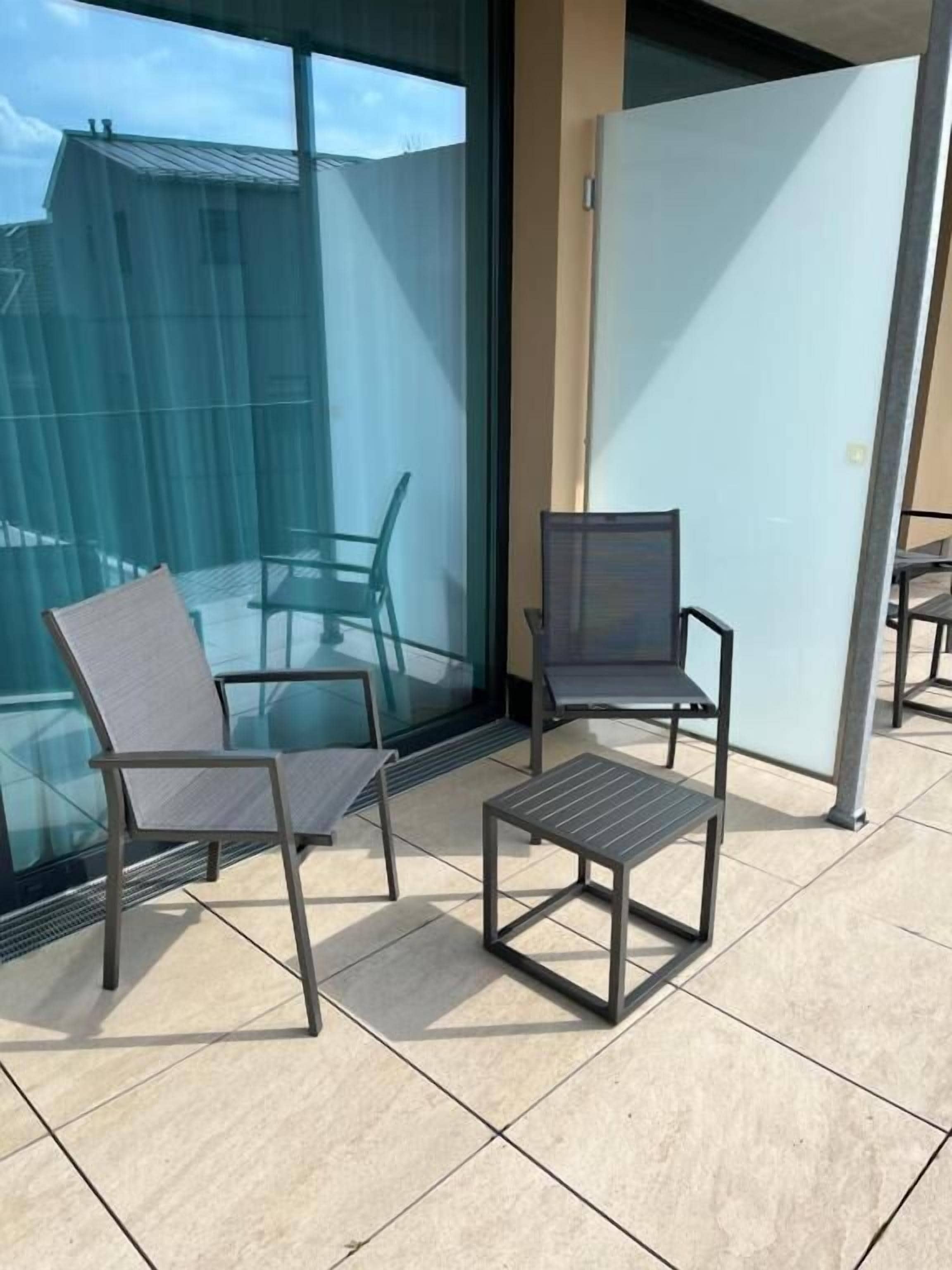 A balcony with two chairs and a small table, large windows, and a view of a building.