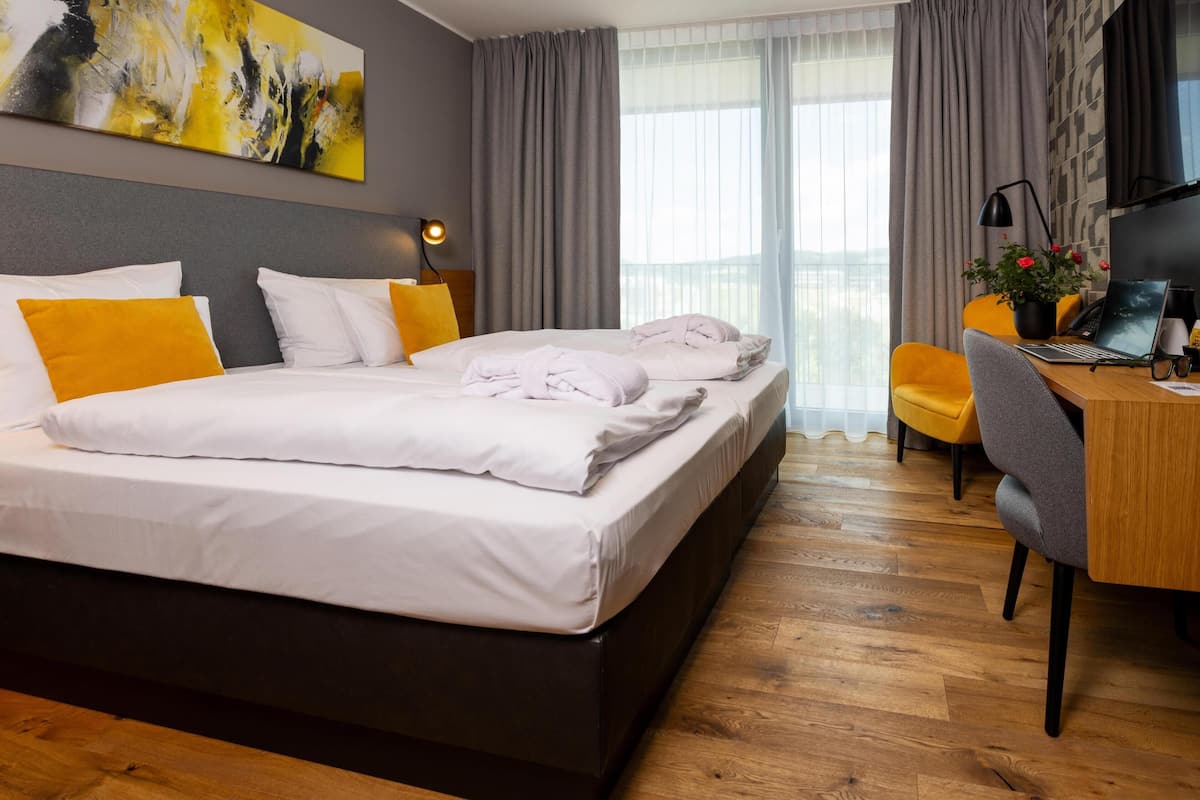 Ein Hotelzimmer mit einem großen Bett, einem Schreibtisch mit einem Laptop, einem Sessel und Blick auf die Stadt durch große Fenster.