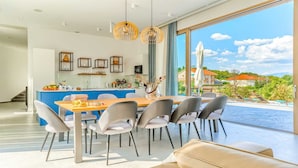 Un comedor moderno con una mesa de madera y sillas grises, una cocina con armarios azules y un amplio ventanal con vistas a una piscina y follaje.