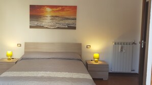 Ein Schlafzimmer mit einem Bett, zwei Nachttischen mit Lampen, einem Wandbild mit Sonnenuntergang über dem Meer und einem Heizkörper.