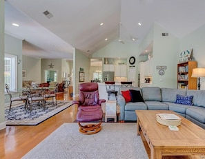 A spacious living room with a wooden coffee table, a blue sofa, and a purple rocking chair. There is a dining area with a glass table and chairs, and a kitchen in the background. - Something for everyone- private pool on the water and pool table! (Hilton Head)