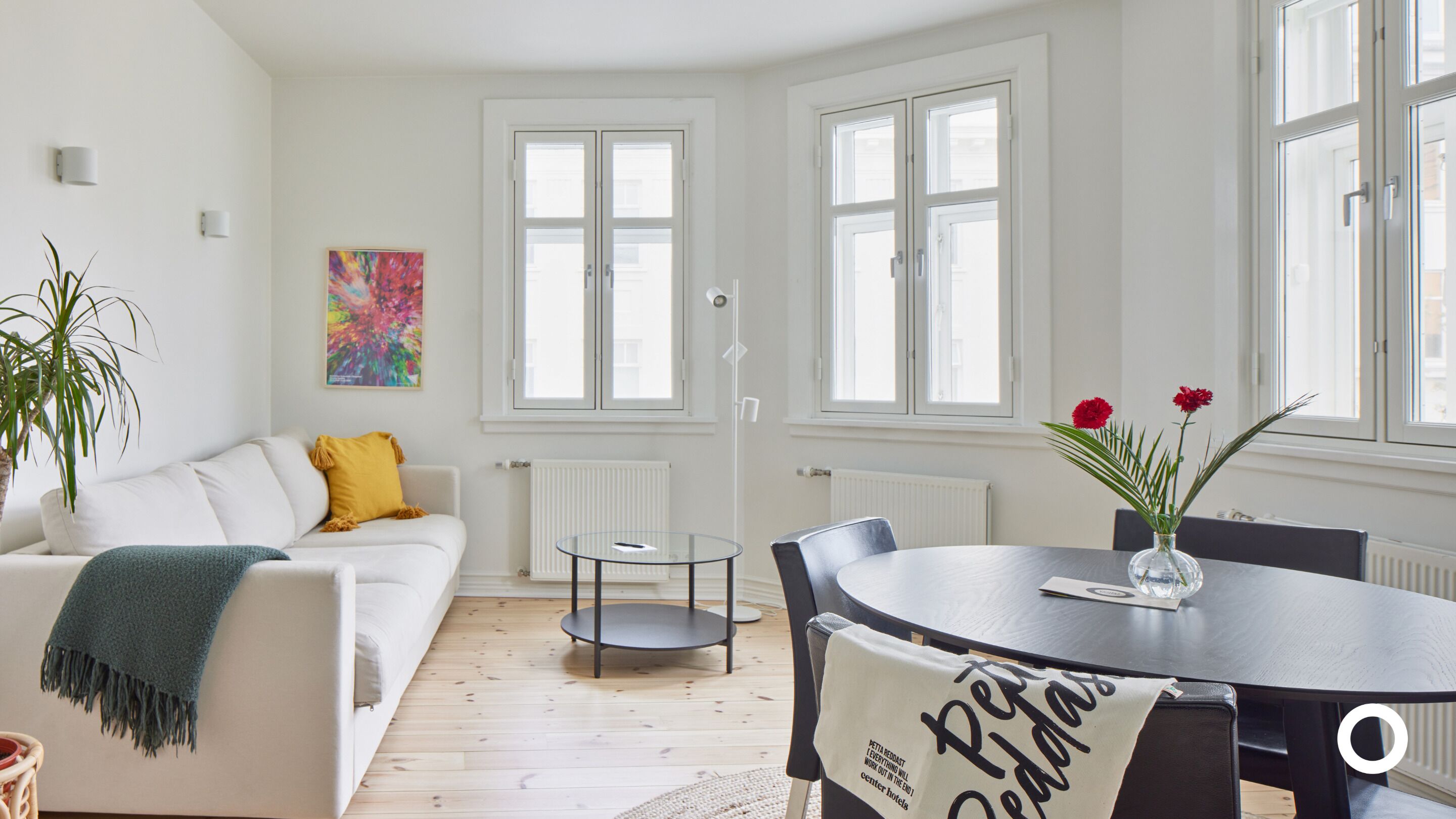 Una sala de estar moderna con un sofá blanco, una mesa de centro de vidrio y una mesa de comedor con sillas. Hay una planta en maceta y un cuadro abstracto y colorido en la pared.