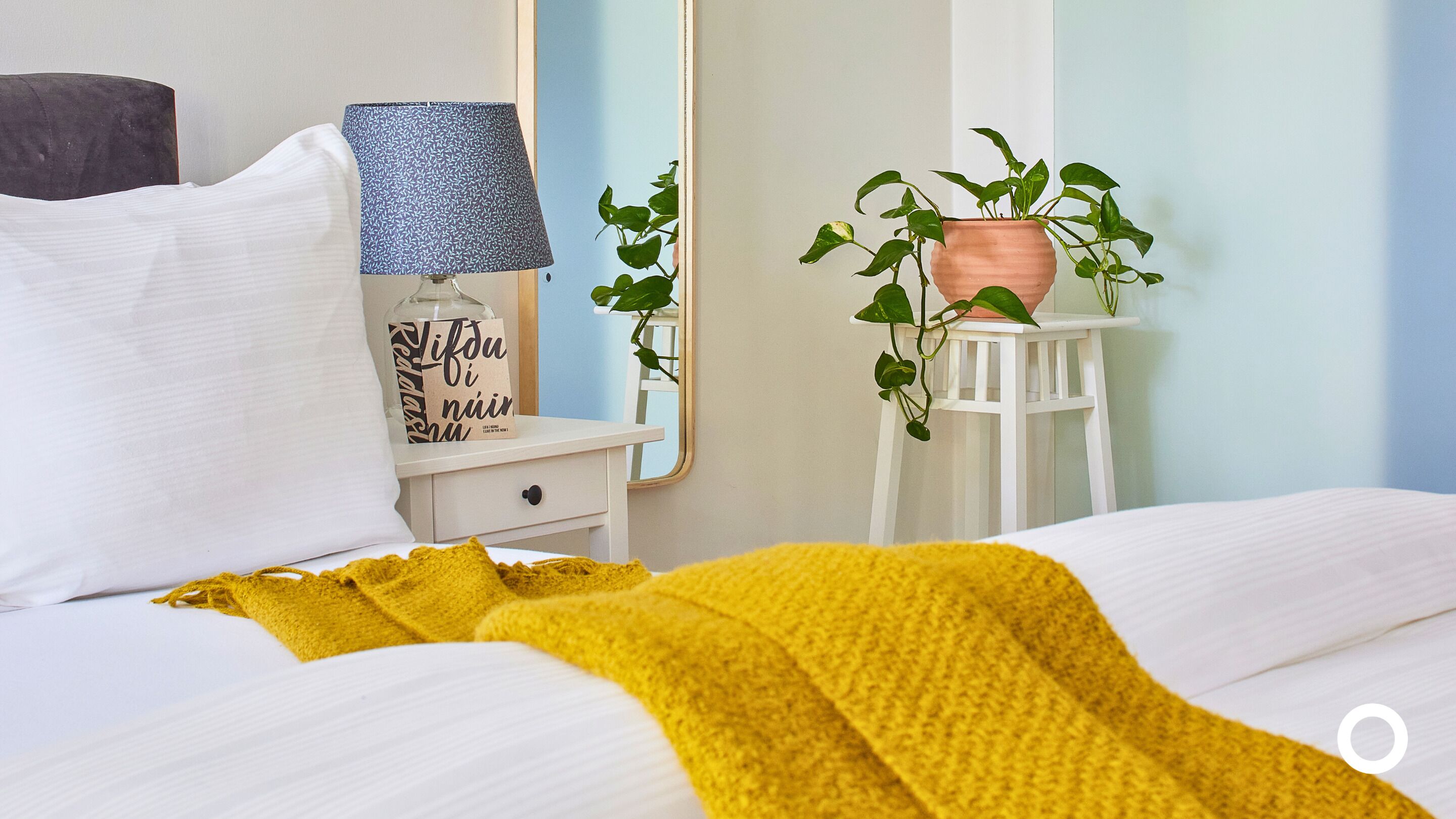 A neatly made bed with a yellow throw blanket, a bedside lamp with a patterned shade, a white nightstand, and a potted plant on a small white stool.