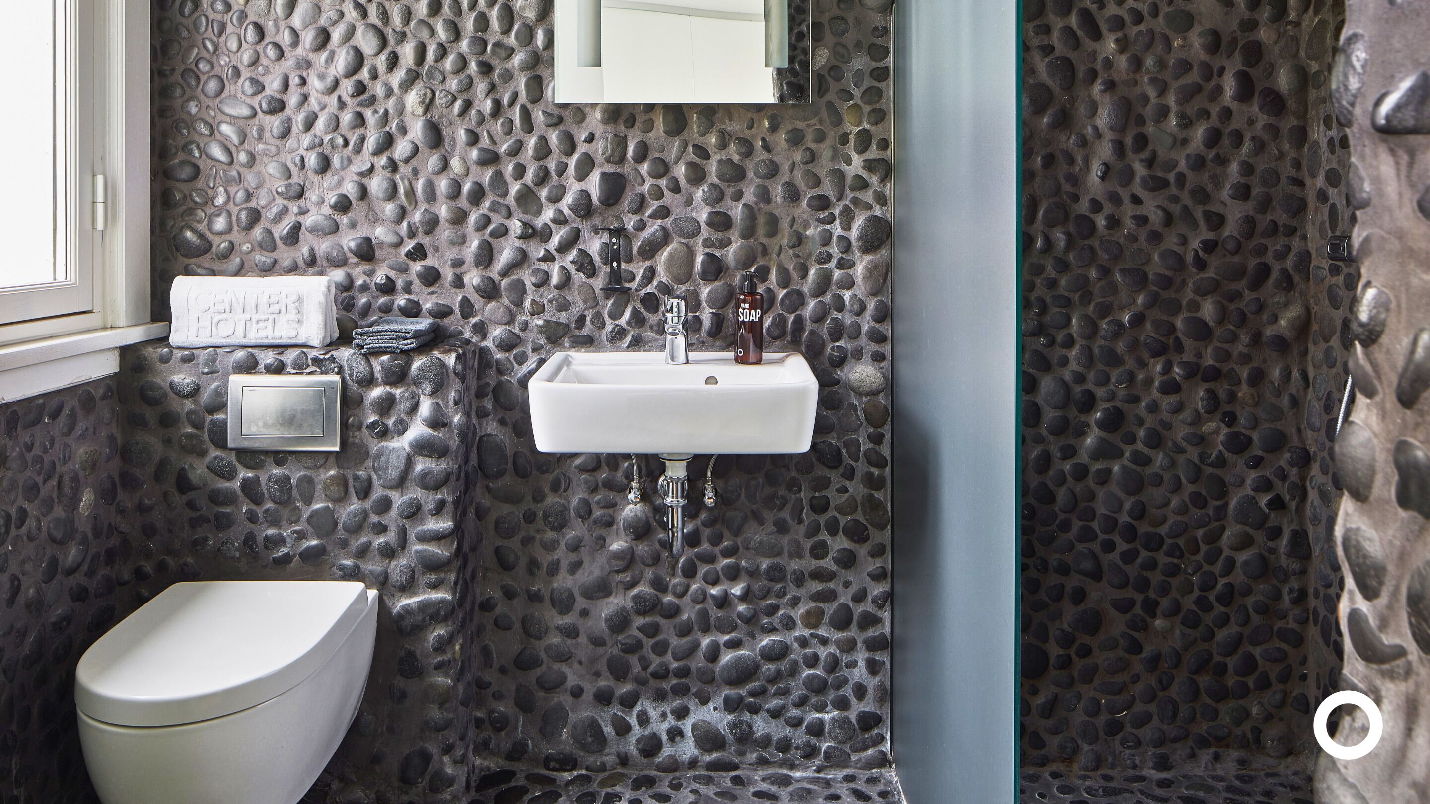 A bathroom with a stone wall, a white sink, a toilet, and a soap dispenser.