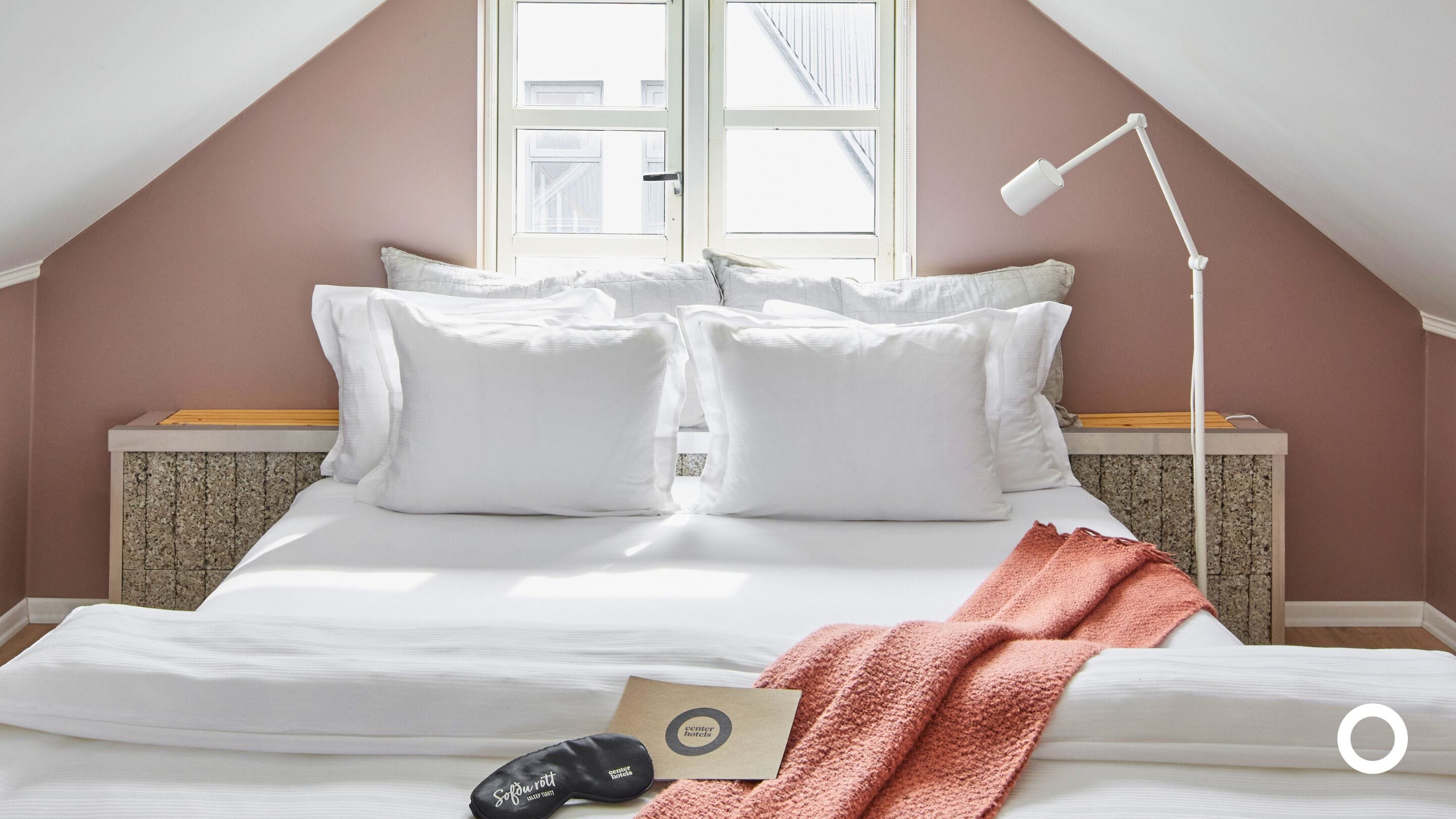 A bedroom with a sloped ceiling, a bed with white bedding, a wooden nightstand, a white desk lamp, and a window with a view of buildings.