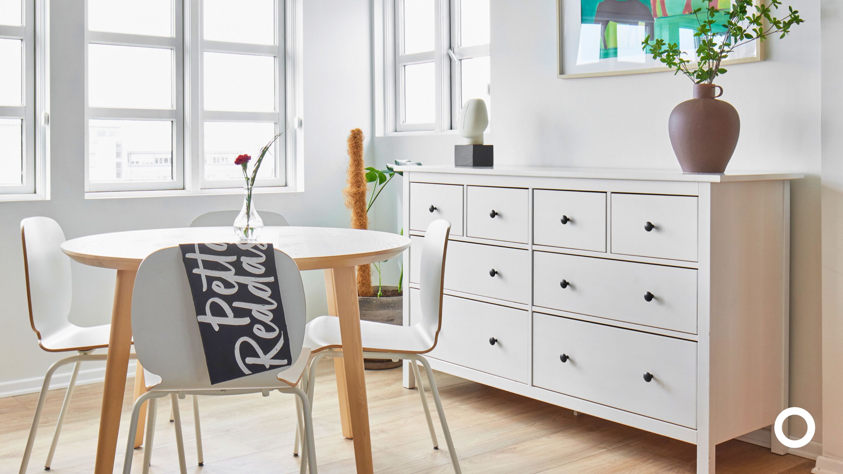 A modern room with a white dresser, a round table with two chairs, and a vase with a plant on the dresser.