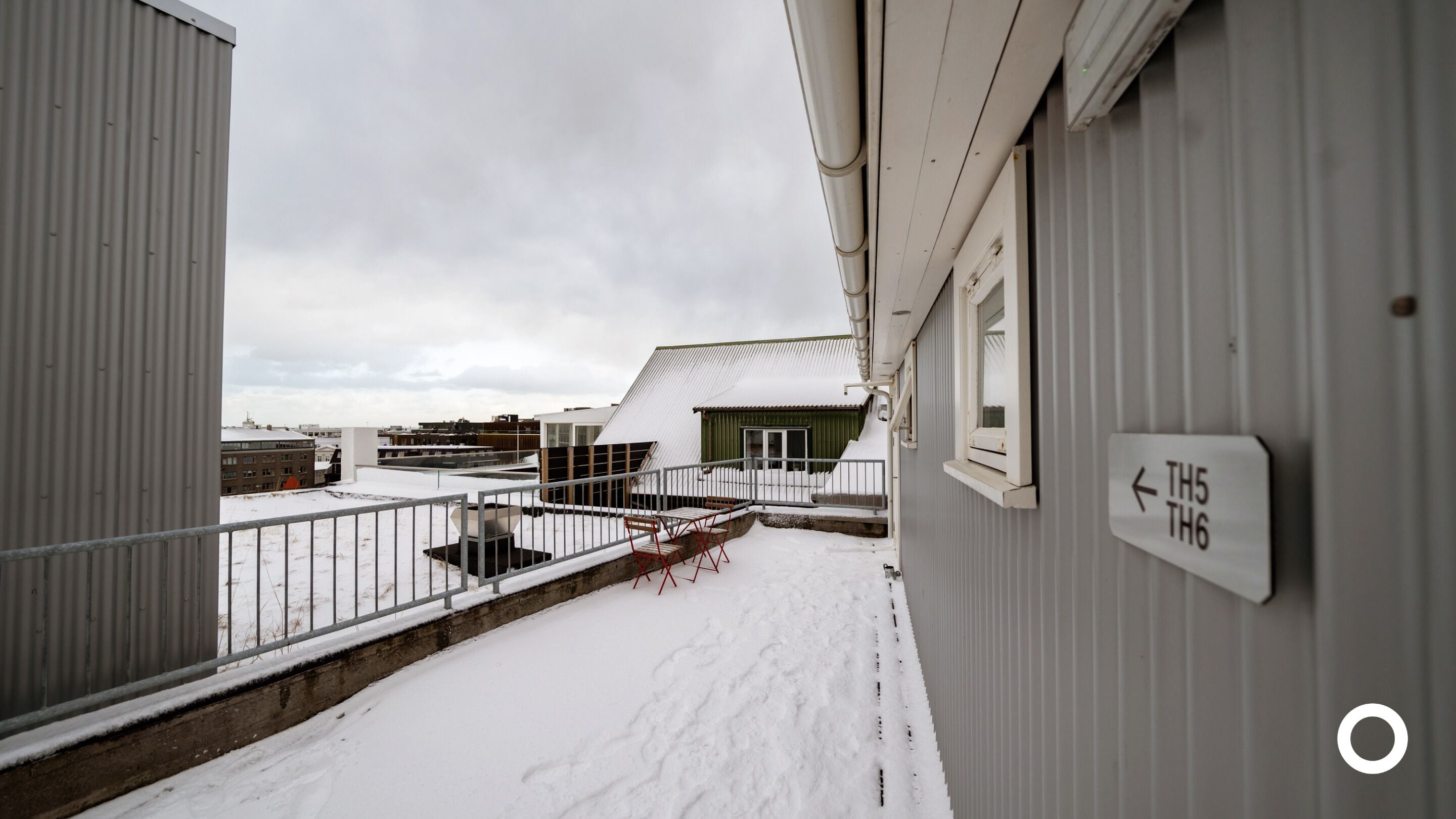 A snow-covered balcony with a metal railing, a sign indicating room numbers, and a view of adjacent buildings.