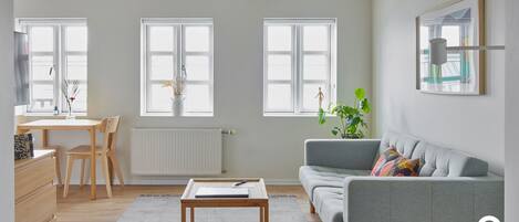 Un salón moderno con un sofá gris, una mesa de centro de madera y un comedor con una mesa y sillas blancas. Tres ventanas amplias proporcionan luz natural.