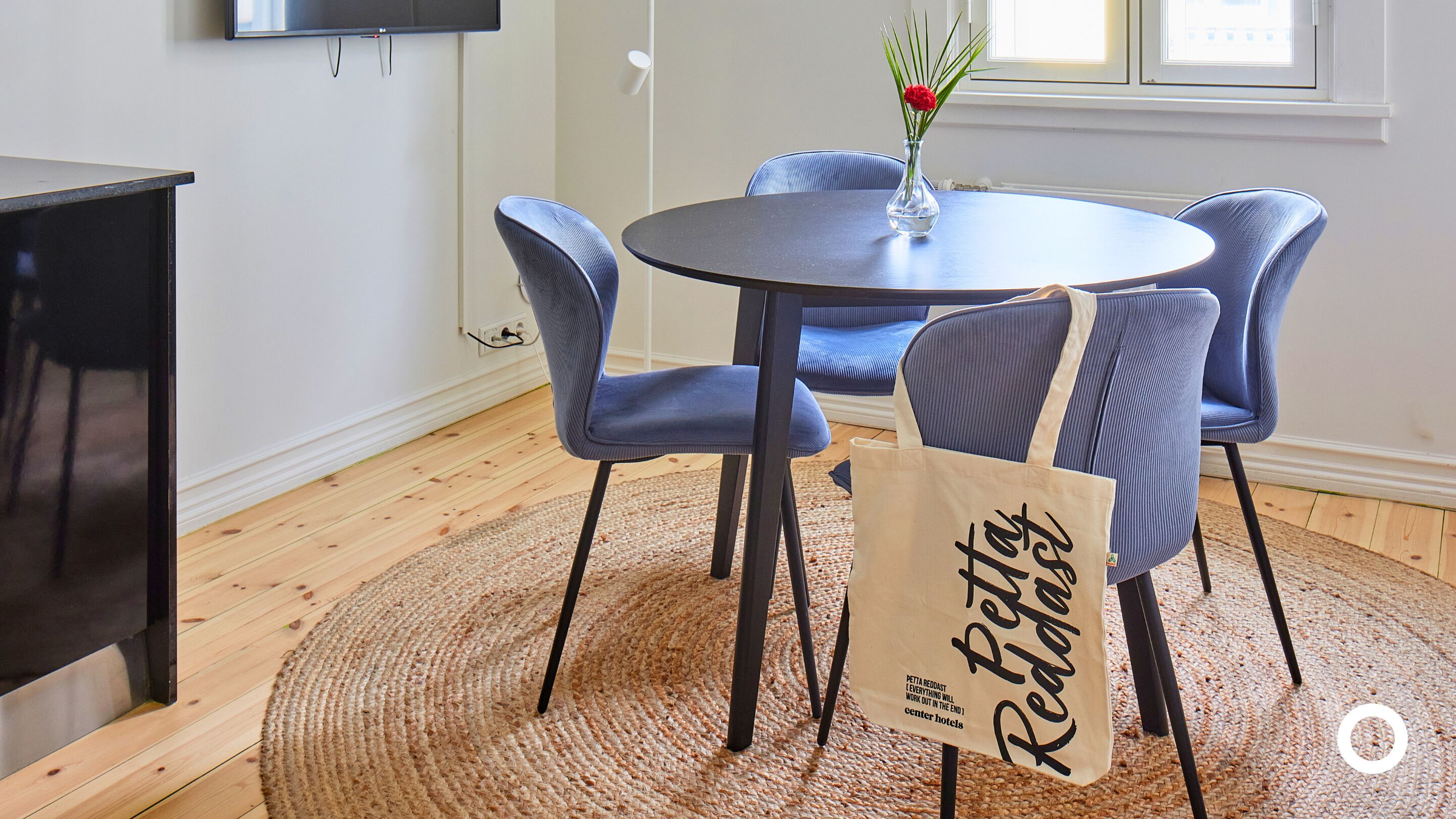 Un comedor con una mesa redonda, cuatro sillas tapizadas en azul, un florero con flores y un bolso de tela en el suelo.