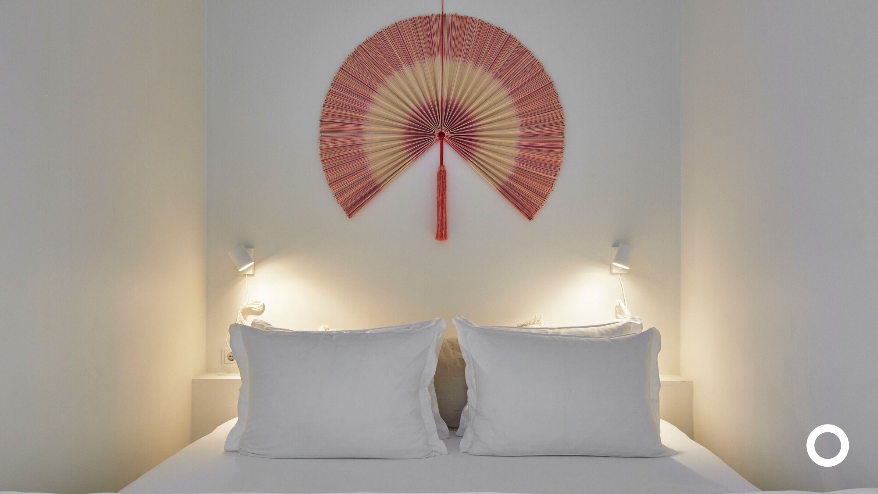 A bed with white linens and two bedside lamps. A red and beige fan is mounted on the wall above the bed.