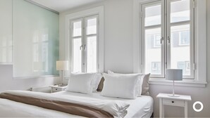 Une chambre à coucher avec un grand lit, deux tables de chevet, une lampe sur pied et une fenêtre donnant sur des immeubles.