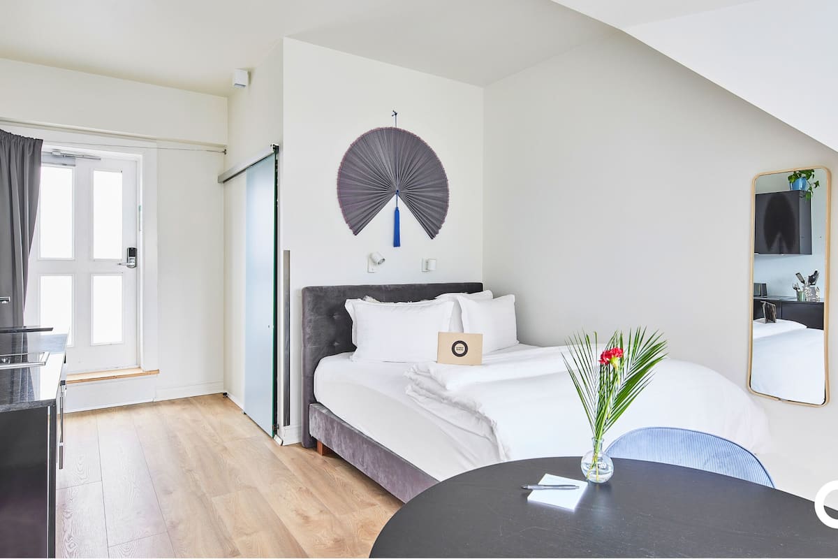 Moderni hotellihuone, jossa on sänky, yöpöytä ja pyöreä pöytä, jonka päällä on maljakko täynnä kukkia.