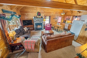 A cozy log cabin interior with a stone fireplace, a television, a dining area, and a kitchen.