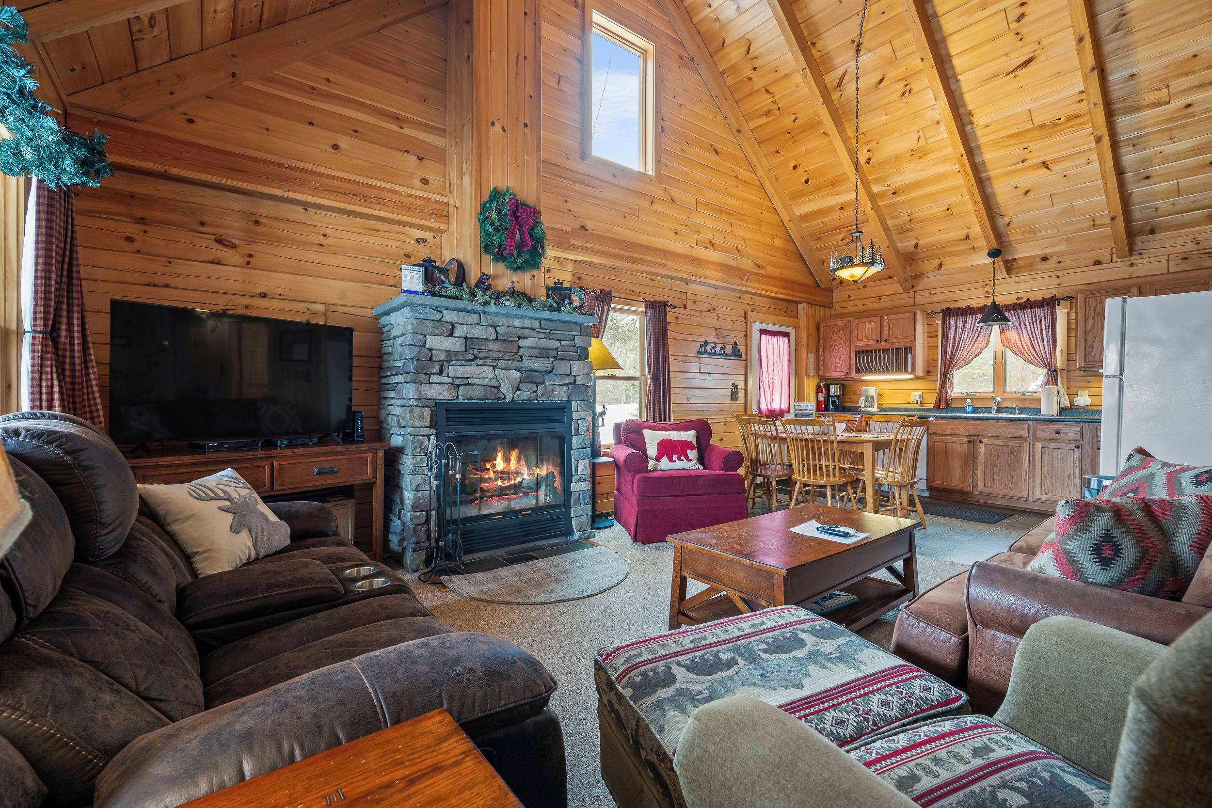 A cozy cabin interior with a stone fireplace, wooden furniture, a television, and a kitchen area.