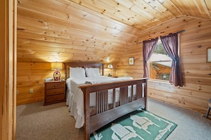 A cozy wooden cabin bedroom with a bed, nightstand, lamp, and a window with curtains.