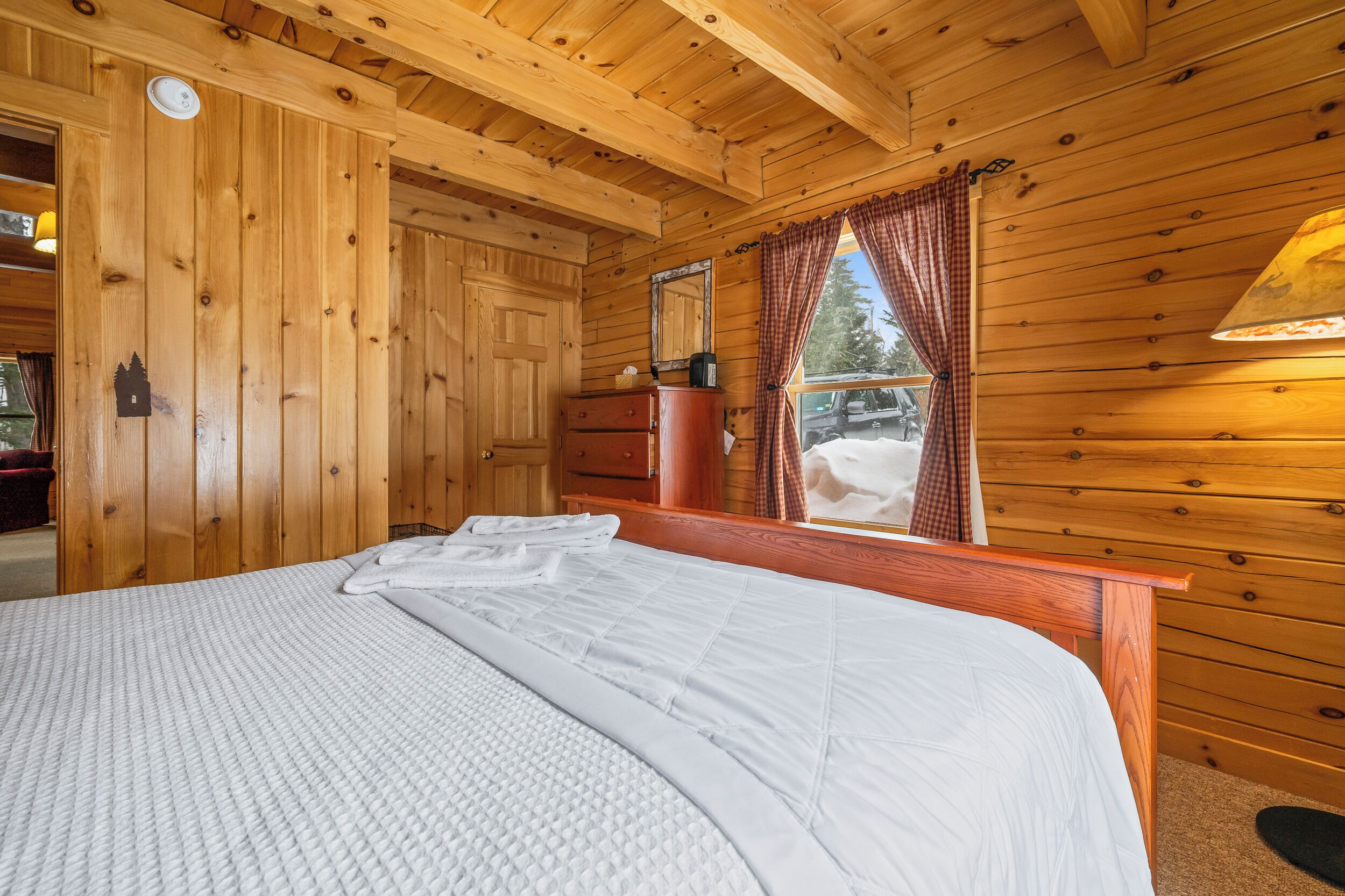 A wooden cabin room with a bed, a dresser, a mirror, and a window with curtains.