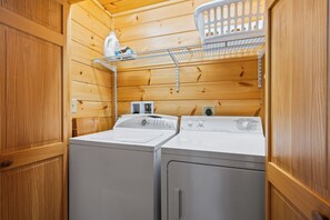 A compact laundry room with two washing machines, a drying rack, and a shelf with a bottle and detergent.