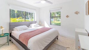 Une chambre moderne avec un grand lit, un ventilateur de plafond et deux fenêtres donnant sur un paysage verdoyant.