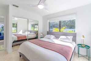 Ein Schlafzimmer mit Bett, Deckenventilator, Fenster mit Blick ins Grüne und einem Nachttisch mit Lampe.