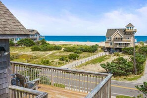 A beachfront property with a wooden deck, a road, and a beach in the background. - Semi-Oceanfront Dog Friendly w/ Saltwater Pool, Hot Tub, Game Room, Volleyball (Avon)