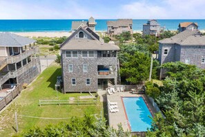 A two-story stone building with a pool, surrounded by greenery and a beach view.