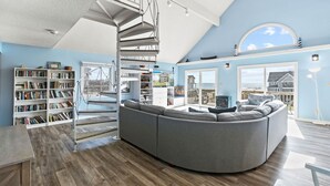 A spacious living room with a curved grey sofa, a spiral staircase, and a large window offering a view of the sea. - Semi-Oceanfront Dog Friendly w/ Saltwater Pool, Hot Tub, Game Room, Volleyball (Avon)
