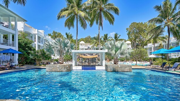 A swimming pool with clear blue water, surrounded by palm trees and white buildings. - 2 Bedroom Oceanfront Penthouse Beach Club (Palm Cove)