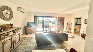 Amplia sala de estar con sofá, una mesa de centro y televisor. Se ve una ventana grande con vista a una piscina y mobiliario exterior.