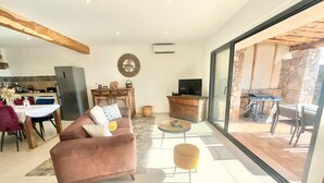 Un amplio espacio de sala con un sofá, una mesa de centro y un televisor. Hay una zona de cocina con una mesa de comedor y sillas. La habitación tiene ventanas grandes y vistas a un patio exterior con una parrilla.
