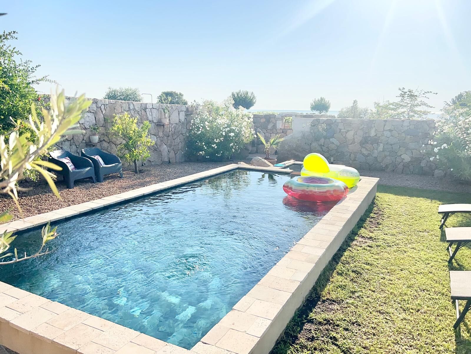 A rectangular swimming pool with a stone border, surrounded by a stone wall and a grassy area.