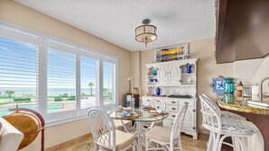 A dining area with a glass table, white chairs, a globe, and a view of the beach. - Direct Beach Views - Sleeps 4, Corner Unit Tennis - Pool (Flagler Beach)