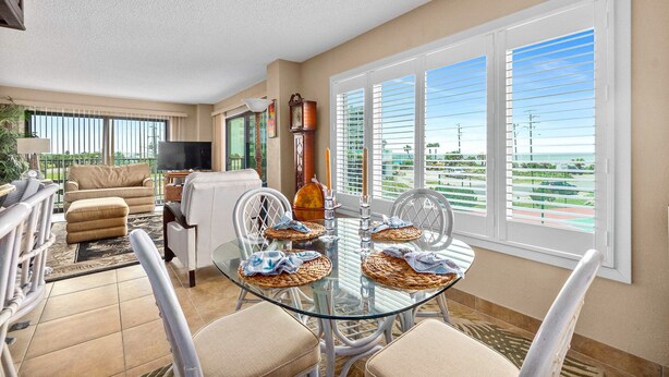 A spacious living area with a glass dining table, white chairs, and a large window with blinds. - Direct Beach Views - Sleeps 4, Corner Unit Tennis - Pool (Flagler Beach)